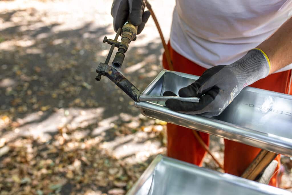 Travail du métal : chalumeau, bac inox, mains gantées Main gantée tenant un chalumeau et insérant une tige métallique dans un bac en acier inox marqué "-D-ENG12". Bricolage extérieur.