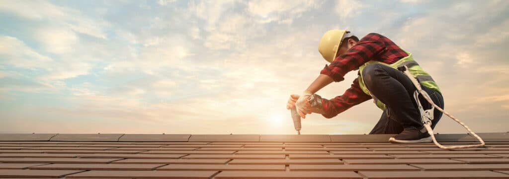 Ouvrier avec casque jaune et gilet de sécurité perçant des tuiles sur un toit au coucher du soleil, équipé d'un harnais.