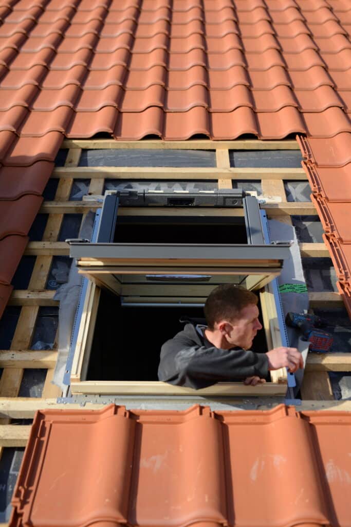 Roofer installant une fenêtre de toit sur un toit en tuiles de terre cuite, avec lattes et membrane visibles.