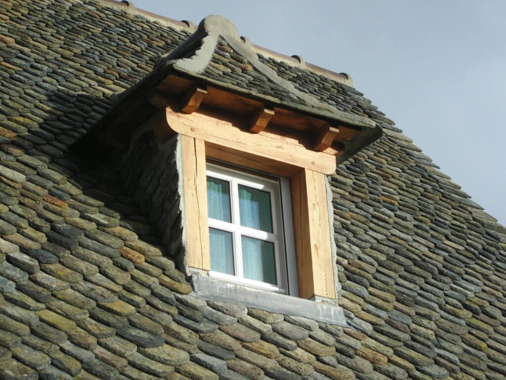 Lucarne en bois sur un toit traditionnel en lauzes foncées, avec une fenêtre à carreaux et habillage en plomb.