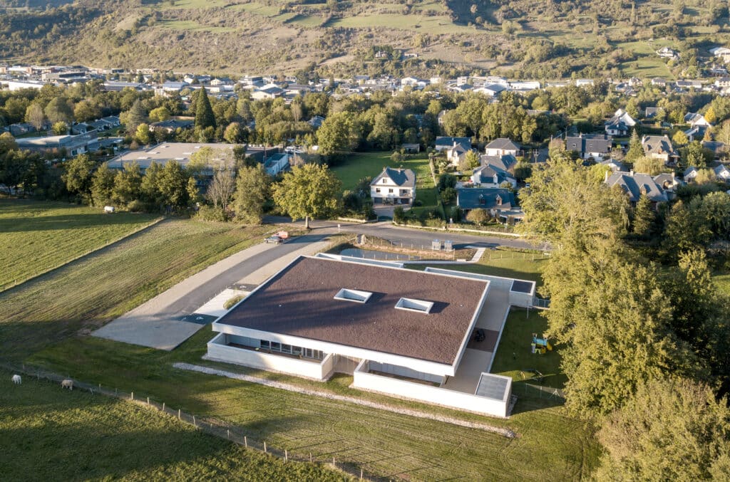 Architecture moderne en montagne, vue aérienne du village. Bâtiment public moderne à toit plat, près de champs verdoyants et d'un lotissement résidentiel. Paysage montagneux. Deux vaches paissent.