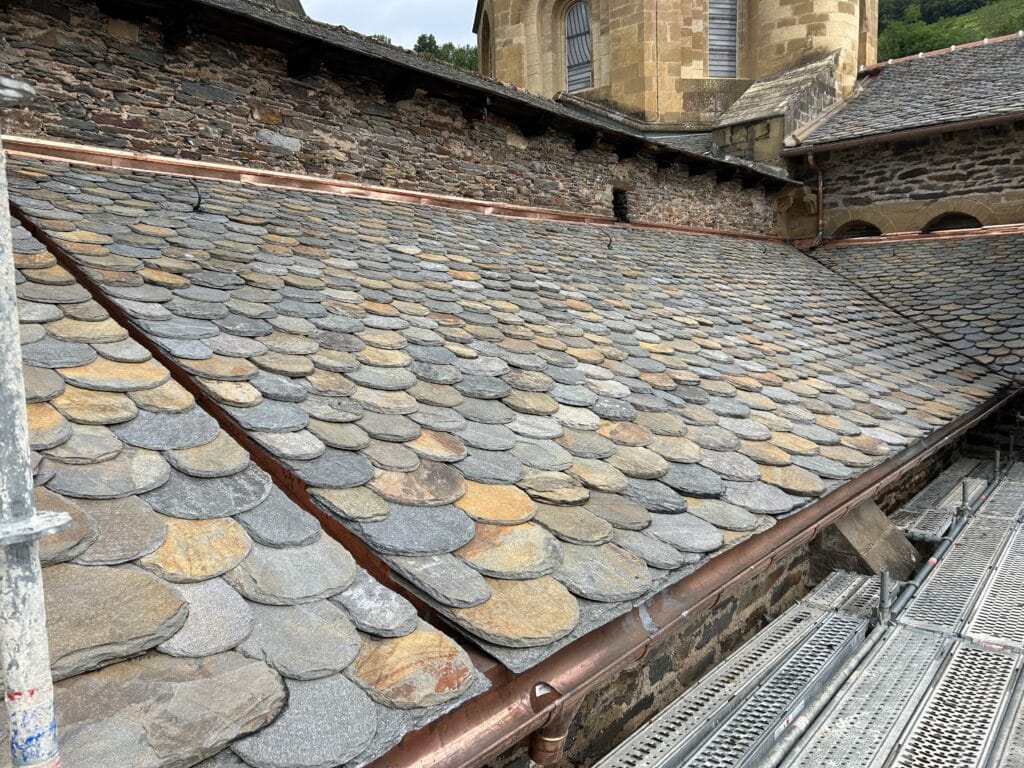 Ardoise multicolore : rénovation de toiture historique. Toit historique restauré en lauzes d'ardoise rondes grises et ocres. Gouttière neuve en cuivre et mur de pierre. Échafaudage de chantier.