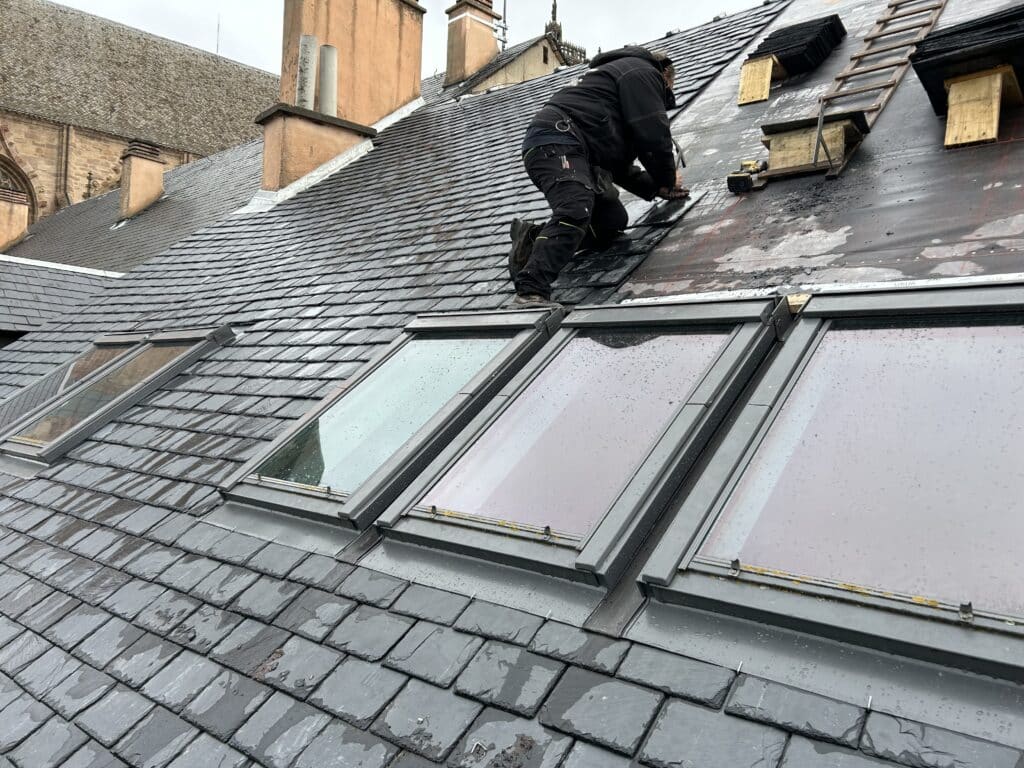 Ardoise : pose professionnelle sur toiture avec lucarnes Couvreur installant des ardoises gris foncé sur un toit. La toiture comporte une série de lucarnes modernes. Bâtiments historiques en arrière-plan.