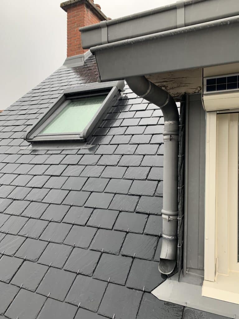 Toiture ardoise : pose, lucarne et gouttière modernes. Toit pentu en ardoise noire mouillée, fenêtre de toit Velux intégrée et descente de gouttière en zinc sur structure de lucarne.