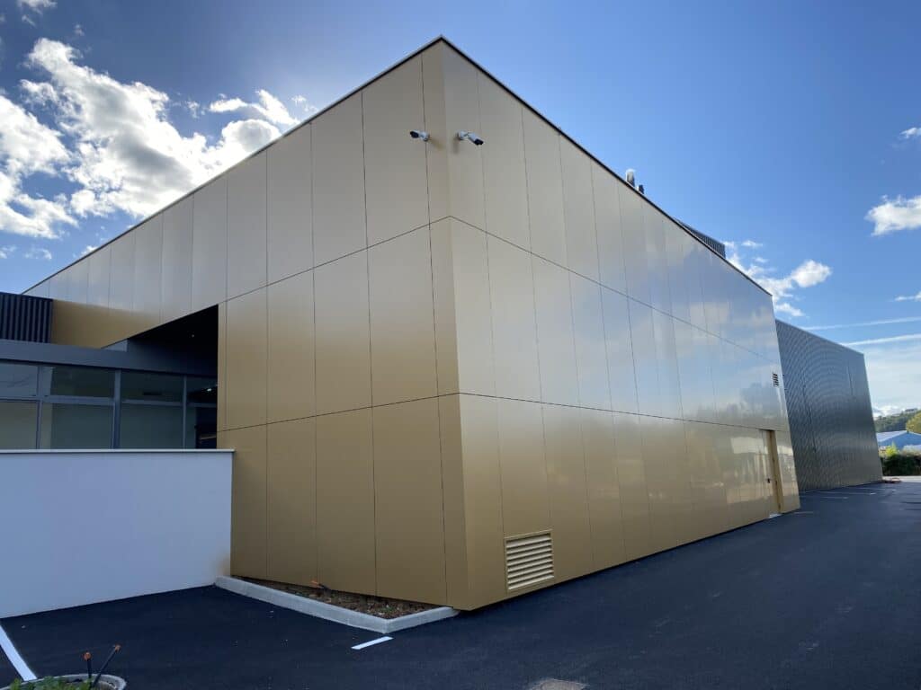 Architecture moderne : Façade en bardage métallique doré Bâtiment moderne à façade dorée sous un ciel bleu, avec caméras de surveillance et parking en asphalte.