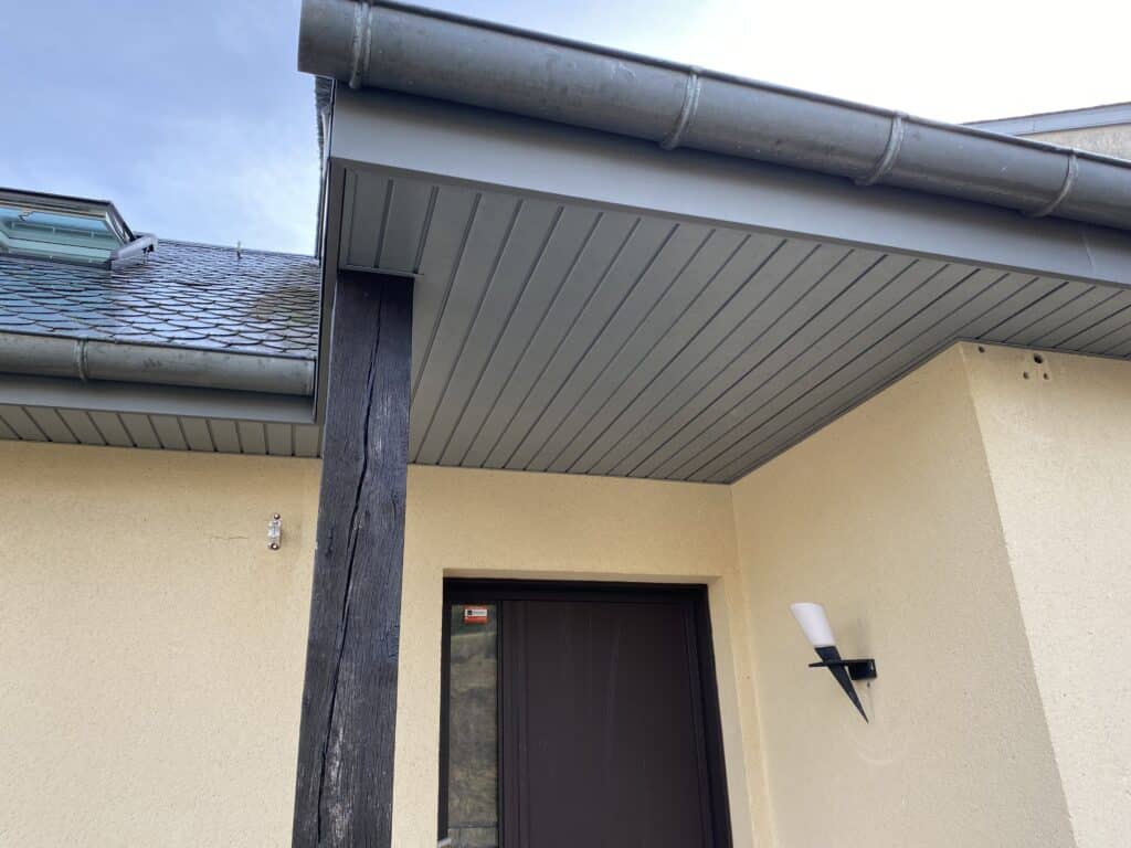 Entrée de maison avec avant-toit gris et poteau en bois Entrée de maison avec auvent gris, pilier en bois sombre, sticker Verisure sur porte foncée et luminaire extérieur.