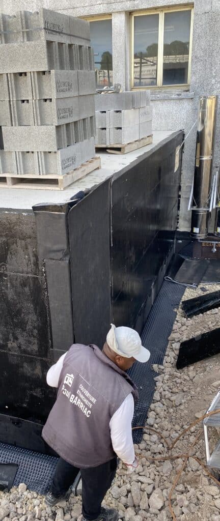 Étanchéité de fondation : pose de membrane protectrice Ouvrier Paul Barriac posant une membrane d'étanchéité Delta-MS sur les fondations d'un chantier de construction.