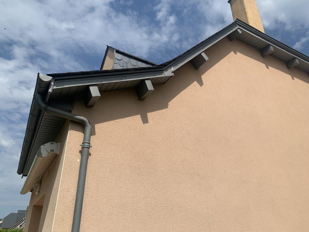 Mur en crépi saumon avec gouttière grise et détails de toiture moderne sous un ciel bleu.