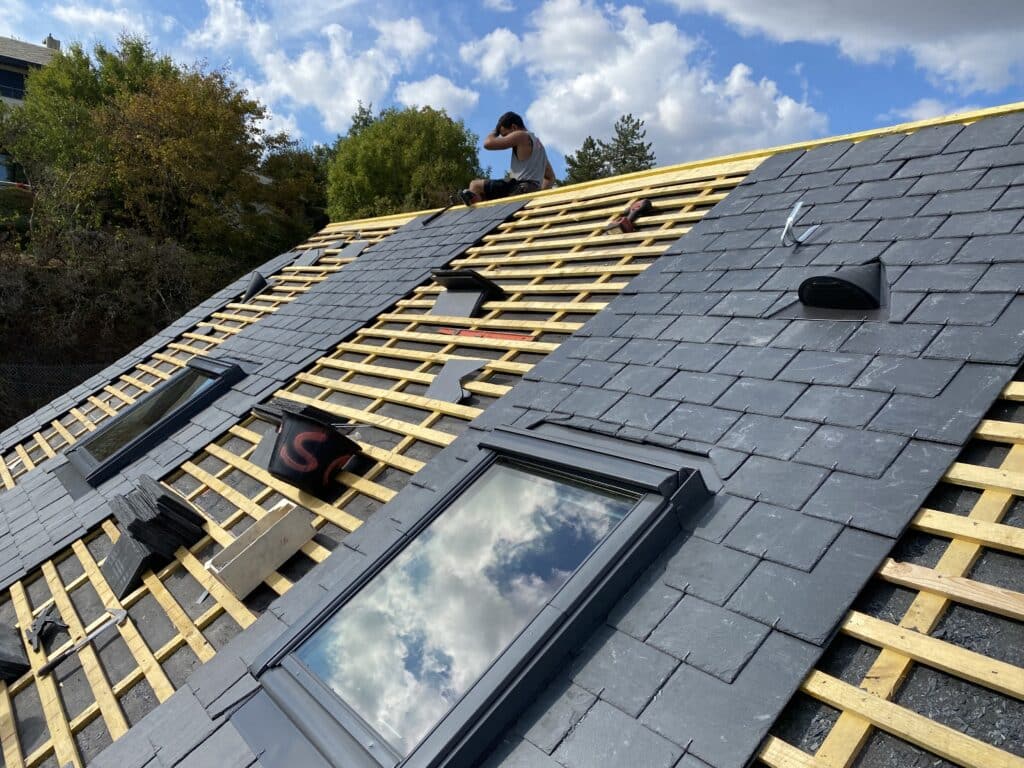 Installation de Toiture en Ardoise avec Fenêtre de Toit Couvreur posant des ardoises sur une charpente en bois avec deux fenêtres de toit sous un ciel bleu nuageux.