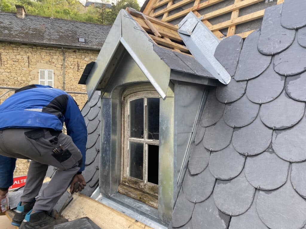 Rénovation de Toiture en Ardoises et Pose de Lucarne Couvreur travaillant sur une toiture en ardoises écailles autour d'une lucarne avec finitions en zinc.