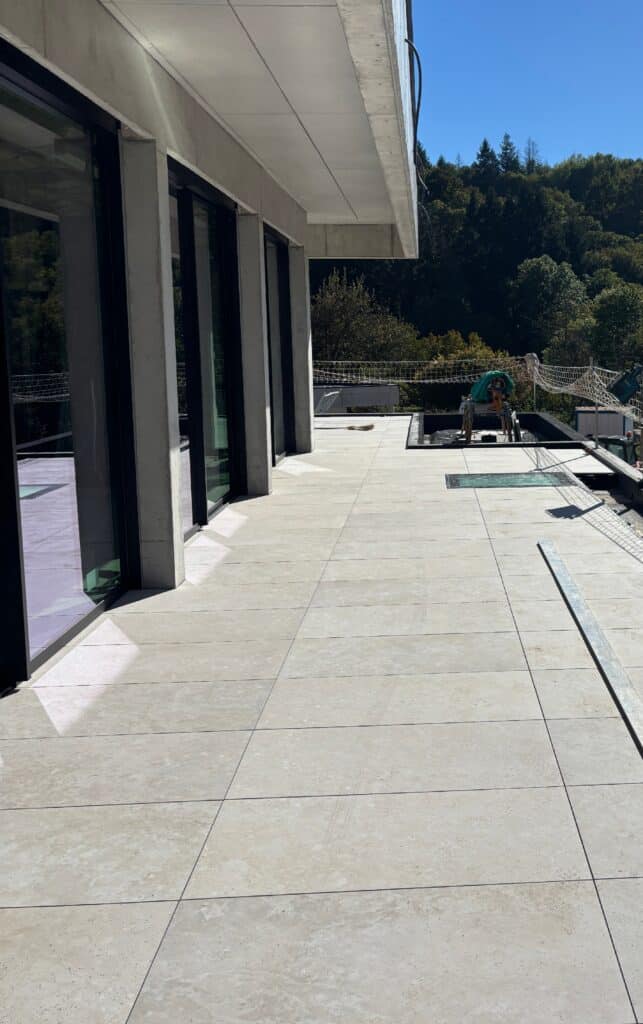 Terrasse moderne en dalles de pierre et béton brut Terrasse moderne avec dalles grises grand format, baies vitrées et vue sur forêt sous un ciel bleu dégagé.