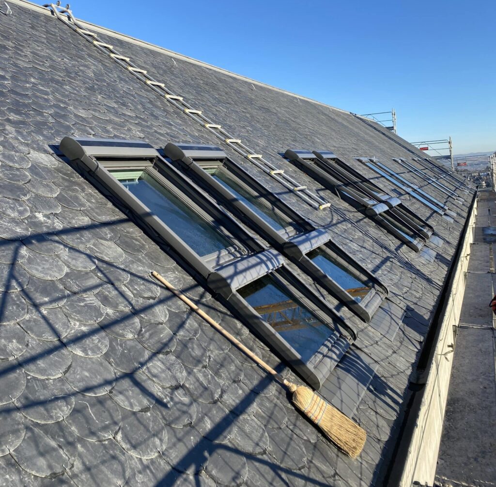 Installation de Fenêtres de Toit sur Toiture en Ardoise Toiture en ardoise avec fenêtres de toit Velux alignées sous un ciel bleu dégagé lors d'un chantier de rénovation.